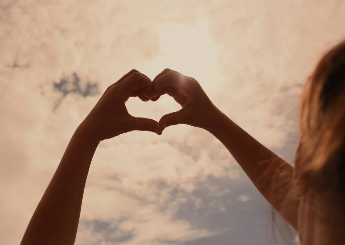 A person is making a heart with their hands against white clouds in the sky.